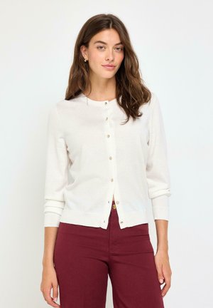Donna con capelli castani lunghi che indossa un cardigan bianco con bottoni e pantaloni bordeaux, in piedi davanti a uno sfondo bianco semplice.