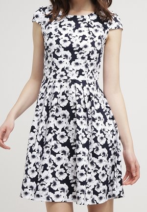 Vrouw die een jurk draagt met korte mouwen, die tot de knie reikt, met een zwart-wit bloemenpatroon en een getailleerde taille.