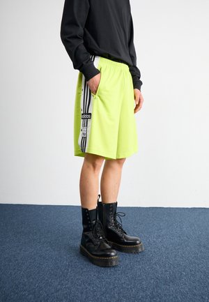 Persona con pantalones cortos Adidas de neón amarillo con rayas negras y blancas y botas de combate negras sobre una alfombra azul frente a una pared blanca.