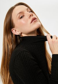 Svart ribbad polotröja med hög krage. Mjuk textur och figurnära design. Modellen håller kragen med ena handen och visar upp ett örhänge.