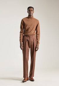 Jersey de cuello alto de lana marrón y pantalones con pliegues. Textura suave, ajuste a medida. Zapatos de cuero marrón completan el conjunto. Fondo neutro.