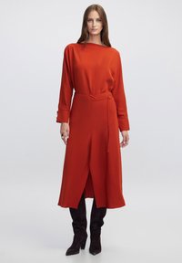 Vestido rojo de manga larga con cintura ajustada y abertura lateral, que presenta una tela suave y una silueta sencilla, combinado con botas negras hasta la rodilla.
