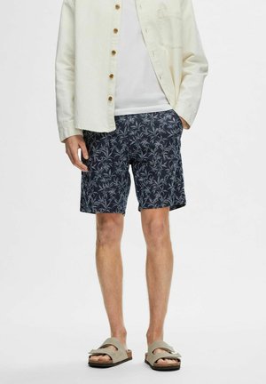 Navy shorts with a white palm leaf pattern, made of lightweight fabric. Paired with a cream button-up shirt and gray sandals.