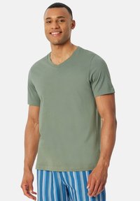 Koszulka T-shirt z dekoltem w serek i krótkimi rękawami w stonowanej zielonej tkaninie bawełnianej, o luźnym kroju i gładkiej fakturze; zestawiona z niebieskimi szortami w paski.