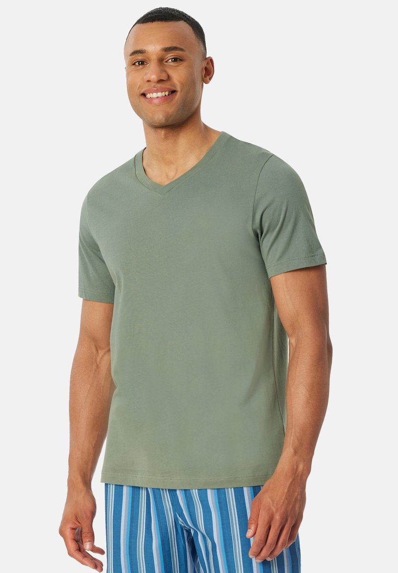 Koszulka T-shirt z dekoltem w serek i krótkimi rękawami w stonowanej zielonej tkaninie bawełnianej, o luźnym kroju i gładkiej fakturze; zestawiona z niebieskimi szortami w paski.