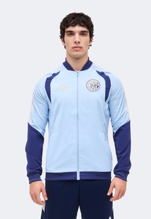 Jeune homme portant une veste de sport zippée bleu clair et bleu marine avec des logos sur la poitrine, debout devant un fond clair uni.