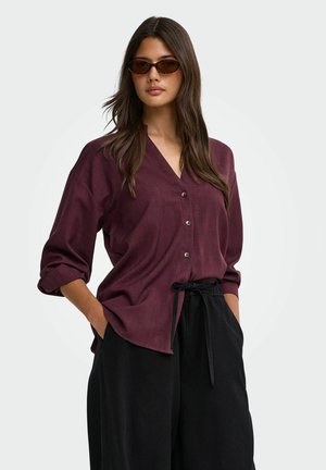 Femme aux longs cheveux foncés portant des lunettes de soleil marron, une chemise ample bordeaux boutonnée avec les manches retroussées et un pantalon noir à cordon avec les mains dans les poches.