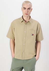 Chemise beige à manches courtes avec un col boutonné, présentant un petit patch logo Tommy Jeans sur la poche poitrine. Tissu en coton doux.