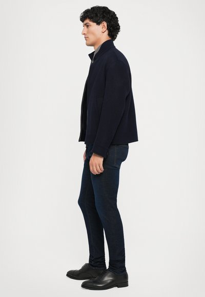 Man standing sideways wearing a navy jacket, dark blue slim jeans, and black leather dress shoes against a plain white background.
