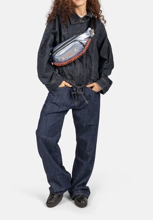 Persoon draagt een losse donkerblauwe spijkerjas en broek met handen in de zakken, en draagt een metallic zilveren schoudertas met oranje en blauwe band.