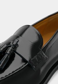 Black patent leather loafers with a decorative tassel and subtle embossed detailing on the band, showcasing a smooth, glossy finish.