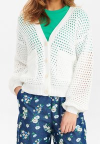 Biały dziergany kardigan z dekoltem w serek, zapinany na guziki, z dwiema kieszeniami. Nosi się go na zieloną koszulę, w połączeniu z granatowymi spodniami w kwiaty.