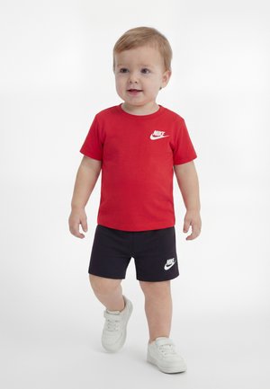 Kisgyermek rövid világosbarna hajjal, vörös Nike pólót, fekete Nike rövidnadrágot és fehér tornacipőt viselve, fehér háttér előtt áll.