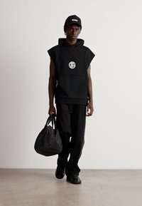 Man wearing black sleeveless hoodie with circular logo, black pants, black cap with "HUGO," and carrying a black duffel bag walking forward.