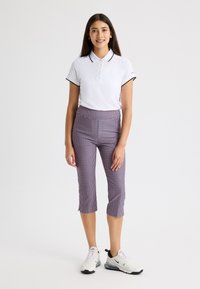 Camisa polo blanca con ribete negro, combinada con pantalones de cuadros morados y blancos hasta la rodilla. El modelo lleva zapatillas deportivas blancas.