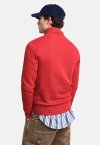 Roter Pullover mit hohem Kragen, aus weichem Stoff. Über einem blau-weiß gestreiften Hemd getragen. Das Modell hat dunkles Haar und eine marineblaue Mütze.