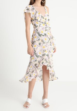 Kobieta ubrana w białą kwiecistą sukienkę kopertową z falbaną na dole i krótkimi rękawkami, w połączeniu z białymi sandałami na obcasie, stoi na jednolitym tle.