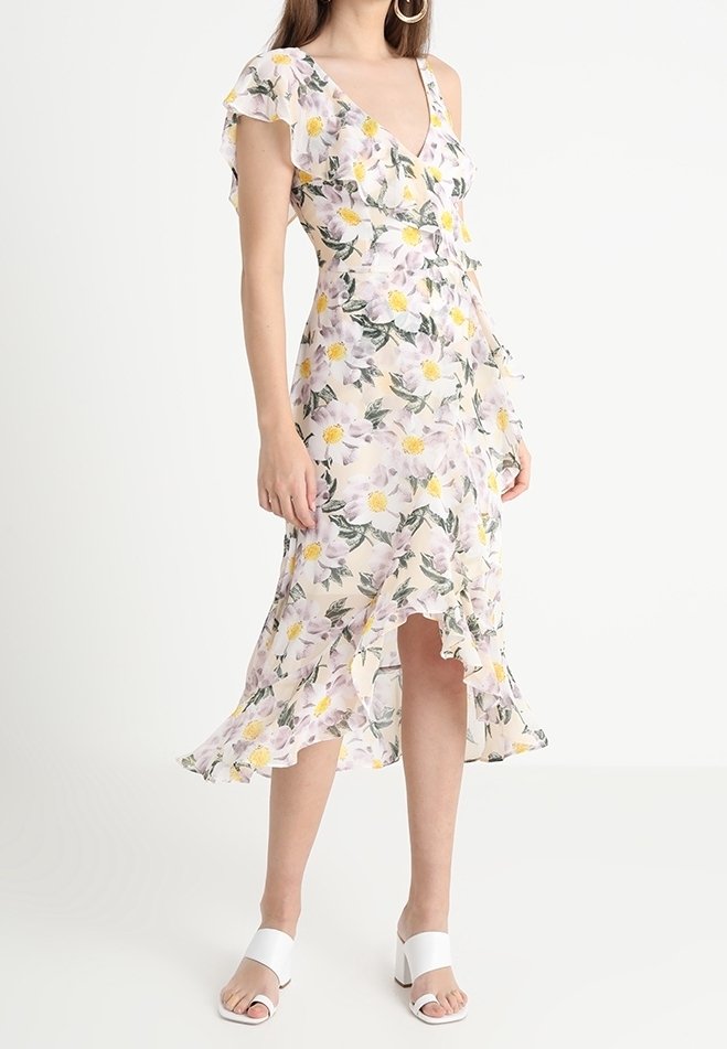 Femme portant une robe portefeuille blanche à fleurs avec ourlet à volants et manches courtes, associée à des sandales blanches à talons, debout sur un fond uni.