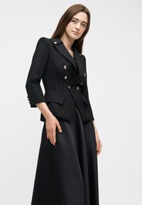 Γυναίκα με εφαρμοστό μαύρο διπλόσαρκο σακάκι με χρυσά κουμπιά και μακριά μαύρη φούστα πάνω σε απλό λευκό φόντο.