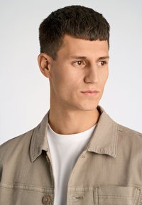 Ljusbeige denimjacka med knäppning, avslappnad passform och enkel krage, bärs över en enfärgad vit skjorta. Slät textur.