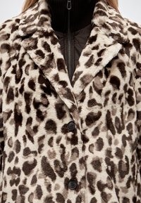 Femme portant un manteau en fausse fourrure à imprimé léopard marron et beige avec de larges revers et des boutons noirs sur un haut noir à fermeture éclair.