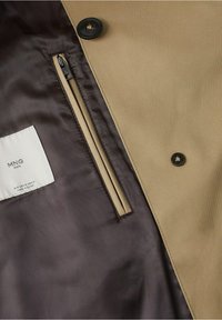 Manteau beige avec boutons noirs partiellement ouvert, révélant une doublure intérieure gris foncé lisse avec une poche verticale zippée et une étiquette de marque blanche.