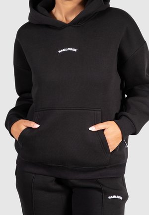 Personne portant un hoodie noir et un pantalon assorti avec des logos blancs "SMILODOX", les mains enfoncées dans la poche avant du hoodie.