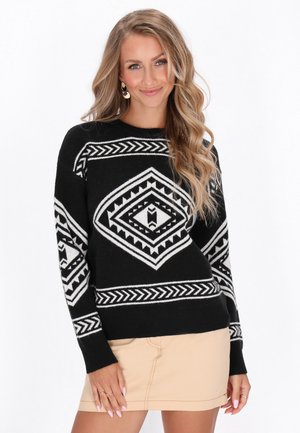 Femme aux longs cheveux blonds porte un pull à motifs noir et blanc et une jupe beige, souriante sur fond blanc.