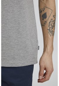 Camiseta de algodón gris con cuello redondo, que presenta una etiqueta lateral, observada en una mano con un tatuaje de cartas de juego, y pantalones oscuros.