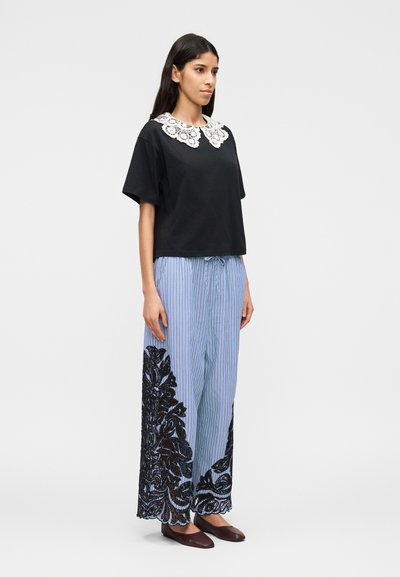 Γυναίκα που φοράει μαύρο τοπ με κοντά μανίκια και λευκή δαντέλα στο γιακά, φαρδιά μπλε παντελόνια με ριγέ και μαύρα λουλουδένια μοτίβα, και σκούρα επίπεδα παπούτσια, όρθια.