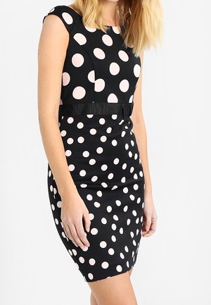 Femme portant une robe noire cintrée avec de gros pois blancs sur le haut et des pois plus petits sur la jupe, debout devant un fond uni.