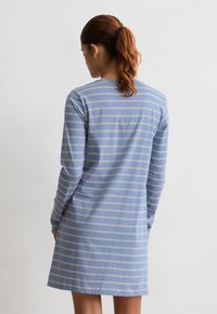 Mujer con cabello castaño en una coleta, vistiendo un vestido de manga larga a rayas horizontales en azul y blanco, de pie contra un fondo blanco liso.
