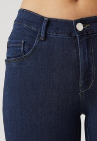 Vaqueros de mezclilla azul oscuro con un ajuste slim. Presentan bolsillos delanteros, un botón plateado y costuras en amarillo contrastante. Textura suave, cintura alta.