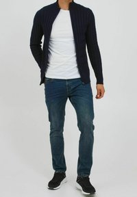 Mann in einem dunkelblauen, mit Reißverschluss versehenen Kabelstrickpullover über einem weißen T-Shirt, blauen Jeans und schwarzen Turnschuhen vor einem schlichten Hintergrund.