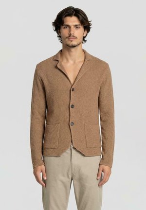 Giovane uomo con capelli scuri che indossa un cardigan marrone testurizzato con bottoni e pantaloni beige, in piedi davanti a uno sfondo chiaro e uniforme.