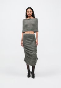 Ženska oblečena v sivo puhasto krajšo zgornjo majico brez naramnic in ujemajočo dolgo krilo, črne legice in črne čevlje na peto, stoji na belem ozadju.