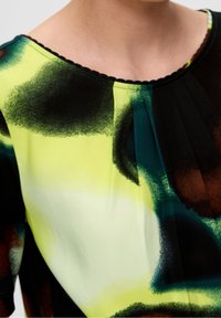 Bluzka z krótkim rękawem uszyta z lekkiego materiału, zdobiona czarno-zielono-żółtym abstrakcyjnym wzorem, z okrągłym dekoltem i plisowanym fasonem.