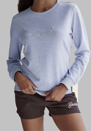 Person trägt ein hellblaues Sweatshirt mit langen Ärmeln und dunkelbraune Shorts, beide mit "Bench"-Logo, die Hände passen den Saum des Sweatshirts an.