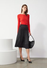 Top rosso a maniche lunghe abbinato a una gonna midi plissettata nera. Tacchi slingback neri e una borsa nera con manico intrecciato.