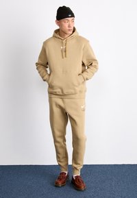 Conjunto de moletom bege composto por hoodie e calças de moletom, feito em mistura de algodão, com um bolso frontal, cordões e o logótipo da Nike em branco. Sapatos castanhos completam o visual.