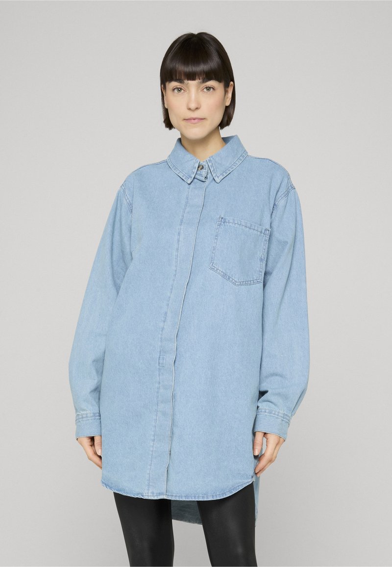 Femme aux cheveux courts et foncés portant une chemise en denim bleu clair oversize avec une poche frontale et des leggings noirs, debout devant un fond uni.