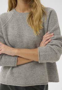 Pull en tricot gris avec une texture côtelée, des poignets larges et un col rond. Le matériau semble doux et confortable, adapté pour un usage décontracté.
