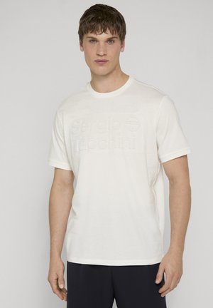 Ung mand med kort brunt hår iført en hvid t-shirt med ophævet Sergio Tacchini-logo og sorte bukser, står mod en ensfarvet baggrund.