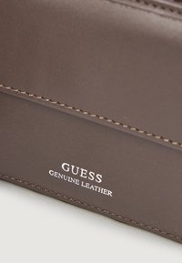 Hnedá kožená peňaženka z pravej kože s prešitými detailmi a embosovanou značkou "GUESS". Hladká textura a minimalistický dizajn.