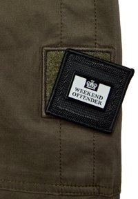 Olívazöld szövet, textúrált folttal. Fekete címke fehér betűkkel "WEEKEND OFFENDER" felirattal, koronás emblémával.