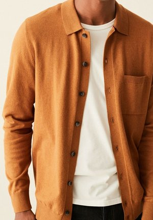 Mann trägt eine kamelfarbene, knöpfbare Strickjacke über einem weißen T-Shirt mit Rundhalsausschnitt, gezeigt vom Hals bis zu den Hüften.