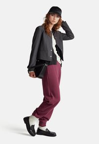 Schwarze kurze Jacke, darunter ein weißes Hemd, kastanienbraune Jogginghose, schwarze Kappe und Loafers mit weißen Socken; Model hält eine strukturierte schwarze Tasche.