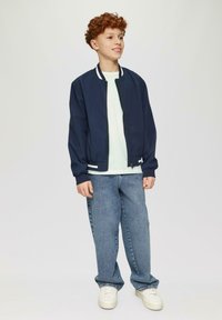 Marine bomberjack met witte accenten, lichtgroen shirt en blauwe broek; model draagt witte sneakers met textuurzolen.