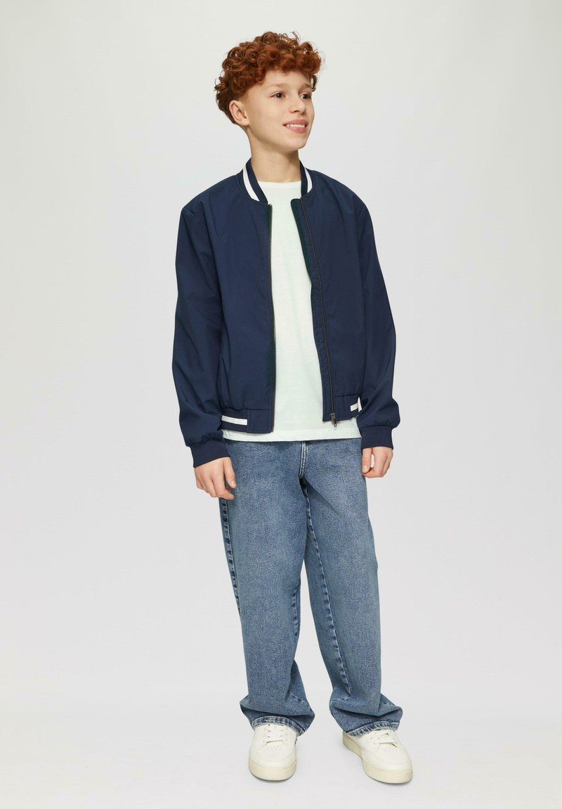 Marine bomberjack met witte accenten, lichtgroen shirt en blauwe broek; model draagt witte sneakers met textuurzolen.