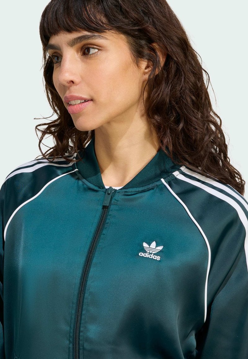 Turkis satin sportjakke med hvide striber og lynlåslukning. Indeholder Adidas-logo på venstre bryst og en opretstående krave.